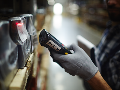 Warehouse management worker holding a barcode scanner standing in front of large pallets of inventory inside a warehouse, standing in the middle of an aisle. 