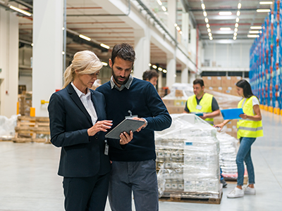 Professional warehouse management designers strategizing over a clipboard with instructions. Background of warehouse workers organizing pallets of inventory in warehouse.